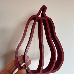 Può includere: Un oggetto tridimensionale cavo a forma di zucca, di colore rosso scuro, con una superficie testurizzata. La struttura è composta da nervature verticali che formano la sagoma di una zucca, con un design interno aperto e un piccolo stelo.