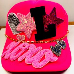 May include: Pink trucker hat with a gold chain, a large pink sparkly "mama" word, a black felt number "1", a pink sparkly star, a silver heart, and a small pink heart with rhinestones.