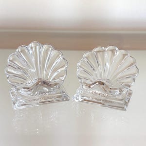 May include: Two clear glass seashell-shaped decorative pieces. Each shell sits atop a square base. The shells have a ridged design, and the bases are also clear. The items are displayed on a reflective surface.