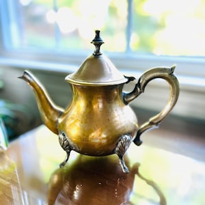 May include: A gold-toned teapot with a rounded body and a lid. The teapot has three decorative feet and a handle that curves up and over the spout.