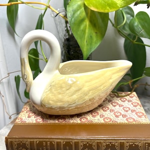 May include: A ceramic swan-shaped gravy boat or planter featuring a glossy cream-colored neck and head with a textured, pale yellow body. The swan has a curved neck and an open, hollow center, showcasing a smooth, glazed finish throughout the piece.