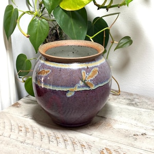 May include: A round ceramic vase with a wide opening and a thick, textured rim. The body features a deep purple base glaze that transitions into a lighter lavender and blue band near the top, accented by small, textured brown leaf-like patterns.