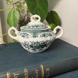 May include: A white and green floral patterned sugar bowl with a lid. The sugar bowl has two handles and a heart-shaped handle on the lid. The sugar bowl is sitting on top of a dark blue book.