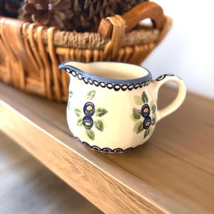 May include: A small, cream-colored ceramic creamer with a blue scalloped rim and handle. The creamer is decorated with a blueberry and leaf pattern. A wicker basket with pinecones is in the background.