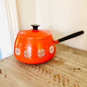 May include: An orange enamel saucepan with a black handle and lid. The saucepan features white decorative circular patterns. The pot is sitting on a light-colored wooden surface.