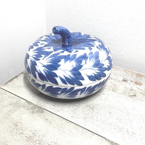 May include: A round ceramic lidded container shaped like a pumpkin, featuring a white base with a hand-painted blue leaf pattern. The lid includes a small, curved blue stem handle at the center, and the overall design is finished in a glossy glaze.