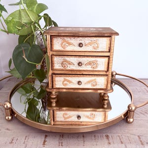 May include: A small, decorative wooden chest of drawers with four drawers. The chest is painted in a distressed style with cream and gold accents. It sits on a gold-toned mirrored tray, reflecting the chest. A green plant is visible in the background.
