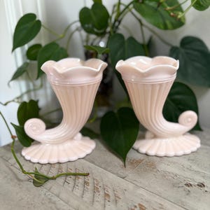 May include: Two pale pink, horn-shaped vases with a ruffled rim and a scrolled base. The vases have vertical ribbed detailing and sit on a scalloped base. The background includes green leafy plants.