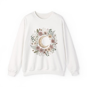 Puede incluir: Sudadera blanca con un diseño de media luna rodeada de una corona floral. La corona incluye flores y follaje rosas, beiges y verdes. El diseño está centrado en la parte delantera de la sudadera de cuello redondo y manga larga.