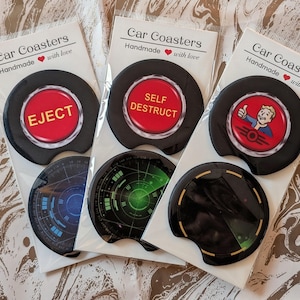May include: Car coasters with different designs. One has a red button with "EJECT", another "SELF DESTRUCT", and a third with a cartoon character. The coasters are black with a circular design, suitable for a car's cup holder.