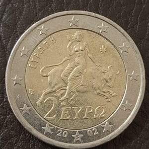 May include: A two-euro coin with a silver outer ring and a gold-colored center. The coin features a depiction of Europa riding a bull, with the text "2 EURO" and the year "2002" visible.