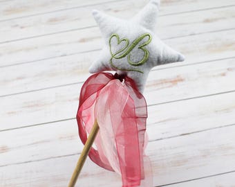 May include: A white star-shaped wand with a pink and white ribbon. The star has a green embroidered letter "B" with two hearts above it.