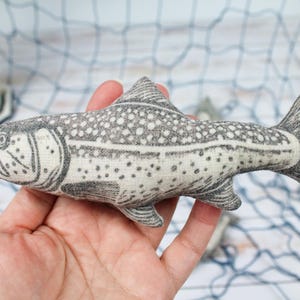 Peut inclure: Une main tient un poisson en tissu gris et blanc avec un imprimé détaillé d'écailles. Le poisson est un jouet en peluche fait main.