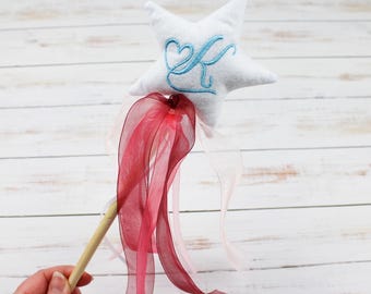 May include: White star-shaped plush wand with a blue embroidered letter "K" and a heart. The wand has a wooden stick and a long red ribbon.