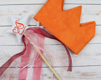 May include: An orange fabric crown and a white star-shaped wand with a wooden handle and flowing ribbons. The star has an embroidered orange letter "S" with heart details. The ribbons are pink and burgundy.