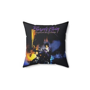 May include: Black square pillow featuring the album art for Prince's "Purple Rain." The design includes the album title in purple script, a motorcycle, and a figure in a purple outfit. The background has a mix of orange, yellow, and blue hues.