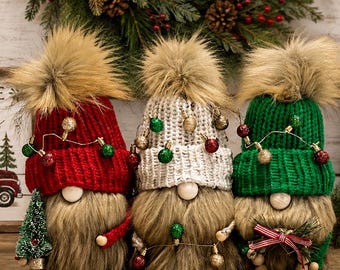 Könnte beinhalten: Drei Weihnachts-Wichtel-Dekorationen mit Kunstpelzbärten und gestrickten Mützen in Rot, Weiß und Grün. Jeder Wichtel hat einen Kunstpelz-Bommel oben. Die Szene umfasst ein Bauernhaus-Weihnachtsschild, einen kleinen Weihnachtsbaum und einen dekorativen Kranz.