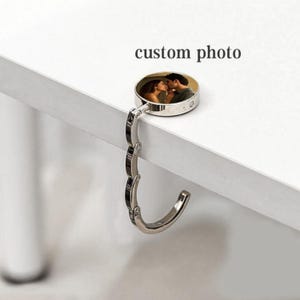 Puede incluir: Un colgador de bolsos de metal plateado con una parte superior circular que muestra una foto personalizada de dos personas frente a frente. El colgador tiene un diseño de gancho plegable de varios segmentos. El texto custom photo está en negro.