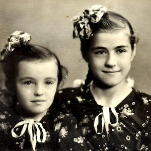 Peut inclure: Photographie en noir et blanc de deux jeunes personnes. Elles portent des robes sombres à motifs floraux et des nœuds blancs au cou. Chacune a un nœud décoratif dans les cheveux.