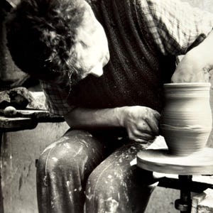 Può includere: Immagine in bianco e nero di una persona che lavora a un tornio da vasaio. La persona indossa una camicia a quadri e un gilet, concentrata sulla modellatura di un vaso di argilla. Il tornio è in movimento.