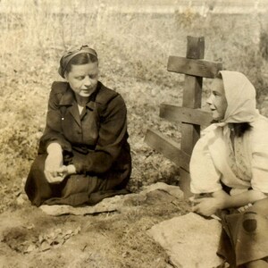 May include: Two women in vintage clothing stand near a wooden cross. The woman on the left is wearing a dark jacket and a headscarf. The woman on the right is wearing a white blouse and a headscarf.