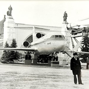 May include: Black and white photograph of a passenger jet on display in front of a large building with statues. The jet has the number "S-42" on its nose. A man in a coat and hat stands in front of the plane.
