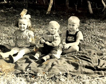 Foto vintage de crianças dos anos 1950 fazendo piquenique ao ar livre. Instantâneo em preto e branco com três crianças pequenas em uma manta. Cena familiar retrô da infância.