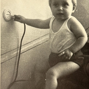 May include: A monochrome photograph showing a young child in a vest and shorts interacting with an electrical socket. The image captures a moment of curiosity, with the child's hand near the plug.