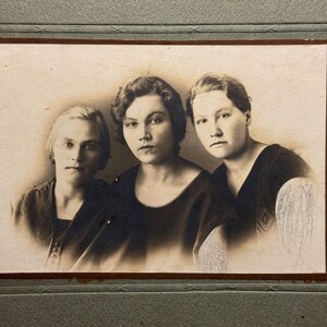 May include: Vintage sepia-toned photograph featuring three women. They are dressed in dark-colored tops, with short hairstyles. The image is framed within a decorative border, suggesting a studio portrait from an earlier era. The photograph is mounted on a textured background.