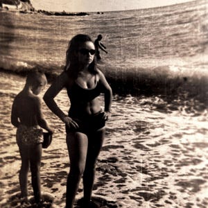 Peut inclure: Photographie en noir et blanc d'une femme et d'un enfant à la plage. La femme porte un maillot de bain noir et des lunettes de soleil, debout dans l'eau. Un enfant se tient à côté d'elle, et une autre personne joue dans les vagues en arrière-plan.