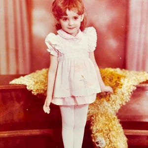 May include: A young girl with red hair in a pink dress and white tights stands next to a large, fluffy teddy bear. The dress has a Peter Pan collar and ruffled sleeves. She wears red Mary Jane shoes. The background is a warm, reddish-brown.