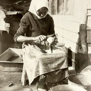 May include: Vintage black and white photograph of a person seated outdoors, peeling a potato. The person wears a headscarf and apron, with metal buckets nearby. The image captures a moment of rural life.