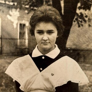 以下が含まれることがあります： 木と建物の前に立っている、黒い襟と白いエプロンが付いた白い制服を着た若い女性の白黒写真です。