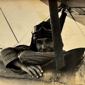 May include: Vintage black and white photograph of a pilot in an open cockpit biplane. The pilot wears a leather helmet and goggles, resting his arms on the cockpit frame. The image has a sepia tone, suggesting an older photograph.