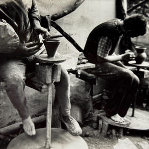 Può includere: Immagine in bianco e nero di due persone che lavorano a torni da vasaio. Una persona modella un vaso di argilla scura, mentre l'altra lavora su un pezzo simile. La scena è rustica, con semplici panche e strumenti di legno.