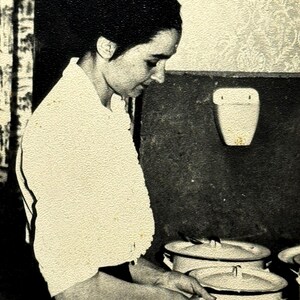 May include: Vintage black and white photograph of a person in a kitchen. The person is wearing a white top and is working with several white pots with lids. The background includes a patterned wall and a wall-mounted fixture.