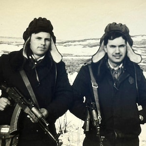 Op de afbeelding: Zwart-witfoto van twee mannen in wintermutsen en donkere jassen, elk met een geweer. Ze staan in een besneeuwde buitenomgeving. De afbeelding is een vintage foto.