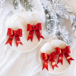 May include: Three pairs of gold hoop earrings with red glitter bow charms. The bows are made of a shiny, sparkly material.