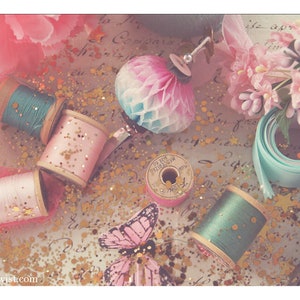 May include: A collection of spools of thread in pink, blue, and white, with gold glitter scattered around them. There is also a pink and blue paper ball ornament, a blue ribbon, and a pink butterfly.