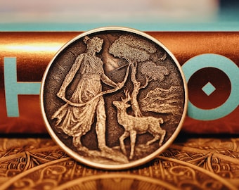 May include: A bronze coin with a raised design of a woman in a flowing robe holding a bow and arrow, standing next to a deer in a forest setting.
