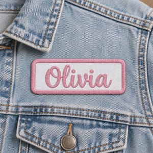 White rectangular patch reading Olivia in pink cursive embroidery with a matching pink stitched border on a faded blue denim jacket, positioned above a buttoned chest pocket.