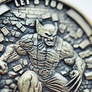 Może przedstawiać: Szczegółowa moneta w kolorze brązu z rzeźbionym wizerunkiem Wolverine'a. Projekt monety zawiera tekst na obrzeżach i tło z rozbitych cegieł, podkreślając motyw superbohatera.