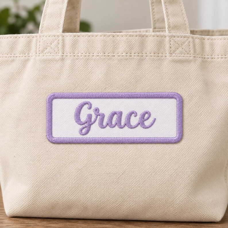 Cream canvas tote with a white rectangular patch reading Grace in purple cursive embroidery and a matching purple stitched border, centered on the front below the handles.