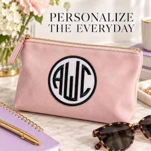 Pink zippered pouch with a white circular patch, Black monogram and matching border, on a tabletop beside a notebook and sunglasses. Heading: Personalize the everyday.