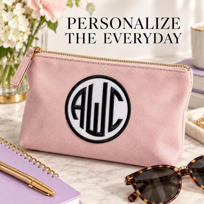 Pink zippered pouch with a white circular patch, Black monogram and matching border, on a tabletop beside a notebook and sunglasses. Heading: Personalize the everyday.