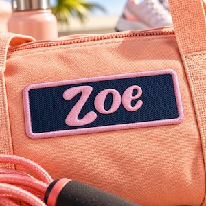 Könnte beinhalten: Eine pfirsichfarbene Sporttasche mit einem rechteckigen dunkelblauen Aufnäher, auf dem der Name Zoe in hellrosa Schreibschrift gestickt ist. Die Tasche hat eine strukturierte Stoffoberfläche und einen passenden pfirsichfarbenen Springseilgriff.