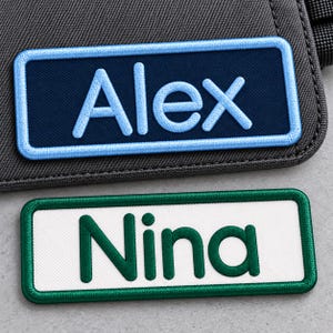 May include: Two rectangular embroidered name patches. The top patch has a dark blue background with light blue text reading Alex and a light blue border. The bottom patch has a white background with dark green text reading Nina and a dark green border.