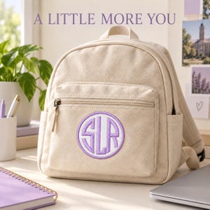 Cream canvas backpack on a desk with a white circular patch and Pale Violet monogram and border on its front pocket. A notebook and laptop sit nearby. Heading: A little more you.