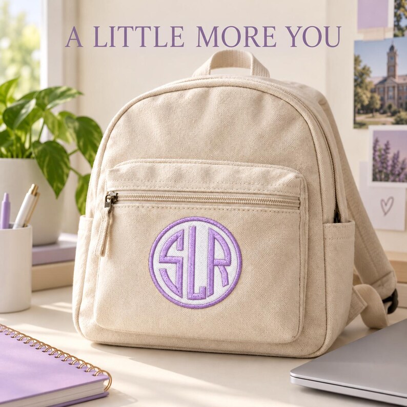 Cream canvas backpack on a desk with a white circular patch and Pale Violet monogram and border on its front pocket. A notebook and laptop sit nearby. Heading: A little more you.