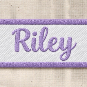 Close view of a complete white twill patch reading Riley in purple cursive embroidery with a purple satin-stitched border, lying on cream woven fabric. Fine diagonal fabric lines and individual thread stitches are visible.
